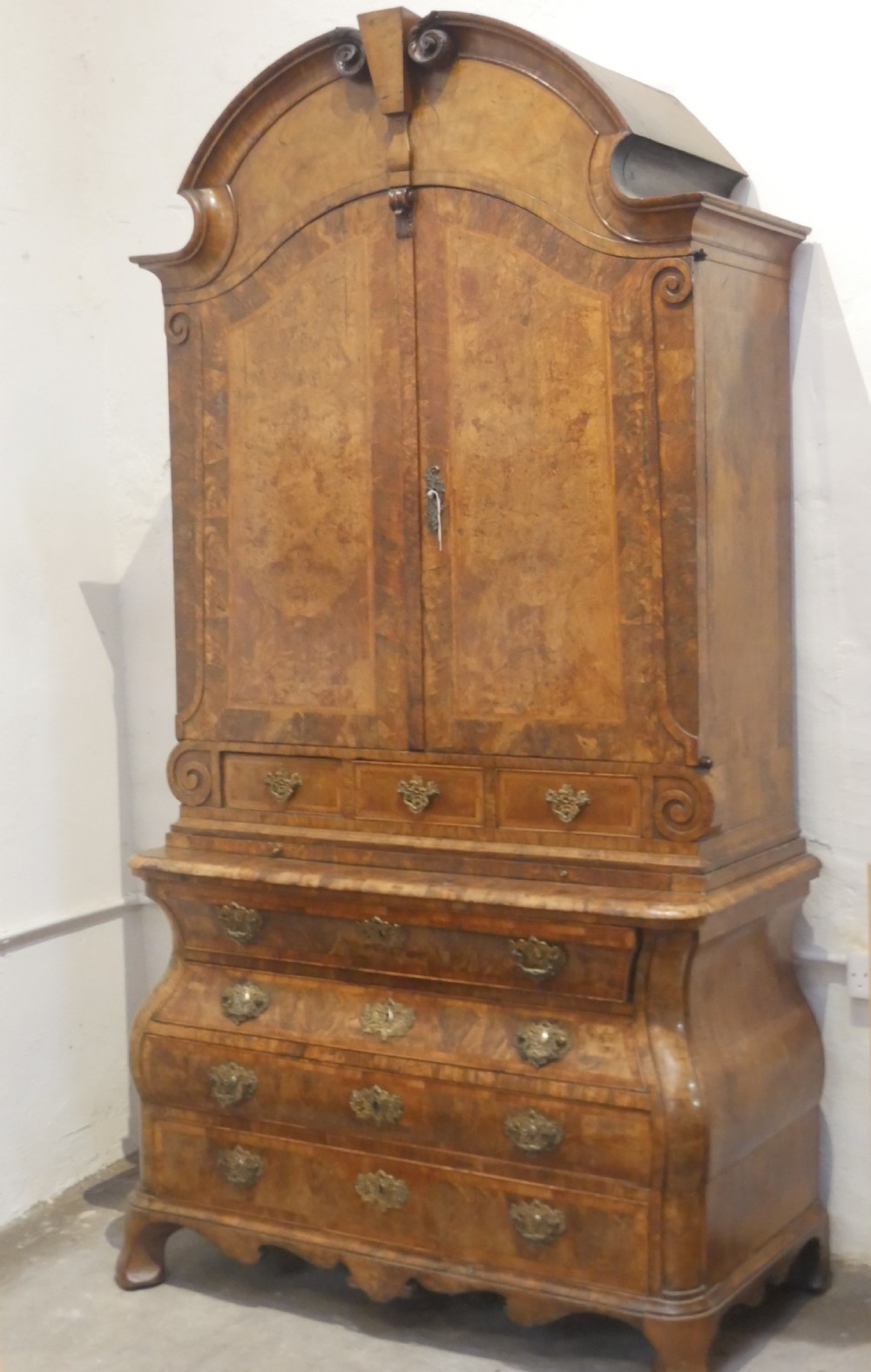 An 18th-century Dutch Rococo burr walnut cabinet featuring an arched moulded cornice, panelled doors, and a bombe three-drawer base with brass hardware.