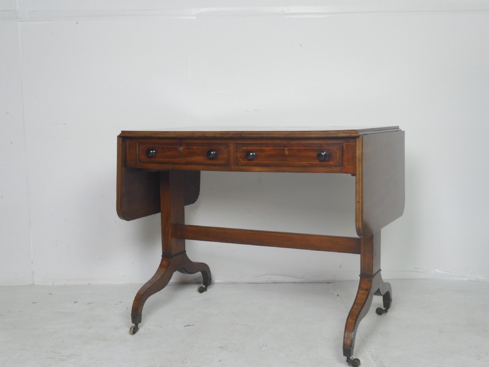 An antique Regency mahogany sofa table featuring two drop leaves, two frieze drawers with wooden knobs, and splayed legs with brass castors.