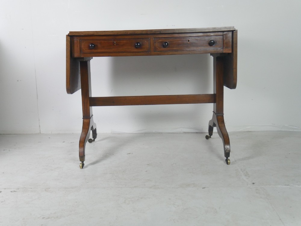 An antique Regency mahogany sofa table featuring two drop leaves, two frieze drawers with wooden knobs, and splayed legs with brass castors. View II