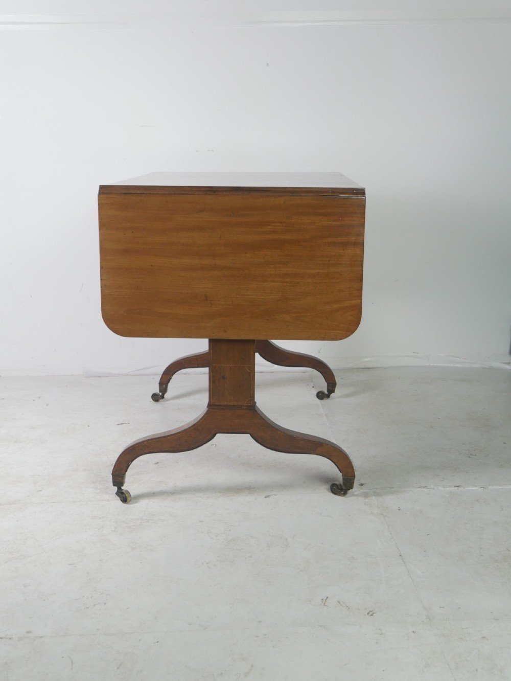 An antique Regency mahogany sofa table featuring two drop leaves, two frieze drawers with wooden knobs, and splayed legs with brass castors. View IV