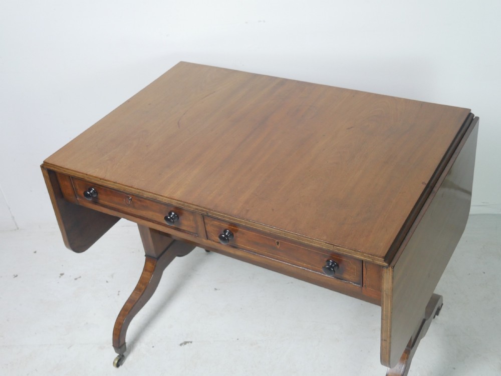 An antique Regency mahogany sofa table featuring two drop leaves, two frieze drawers with wooden knobs, and splayed legs with brass castors. View V