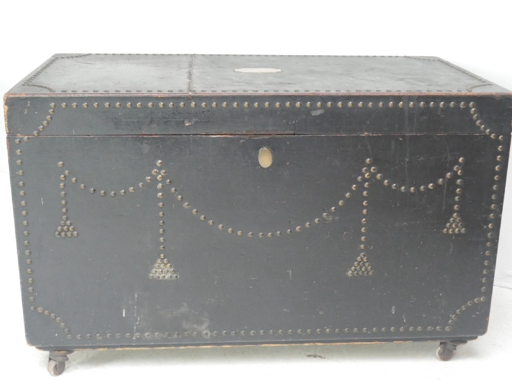A black leather-covered storage trunk adorned with brass studding in festoon drape patterns, featuring side handles and castors. View V