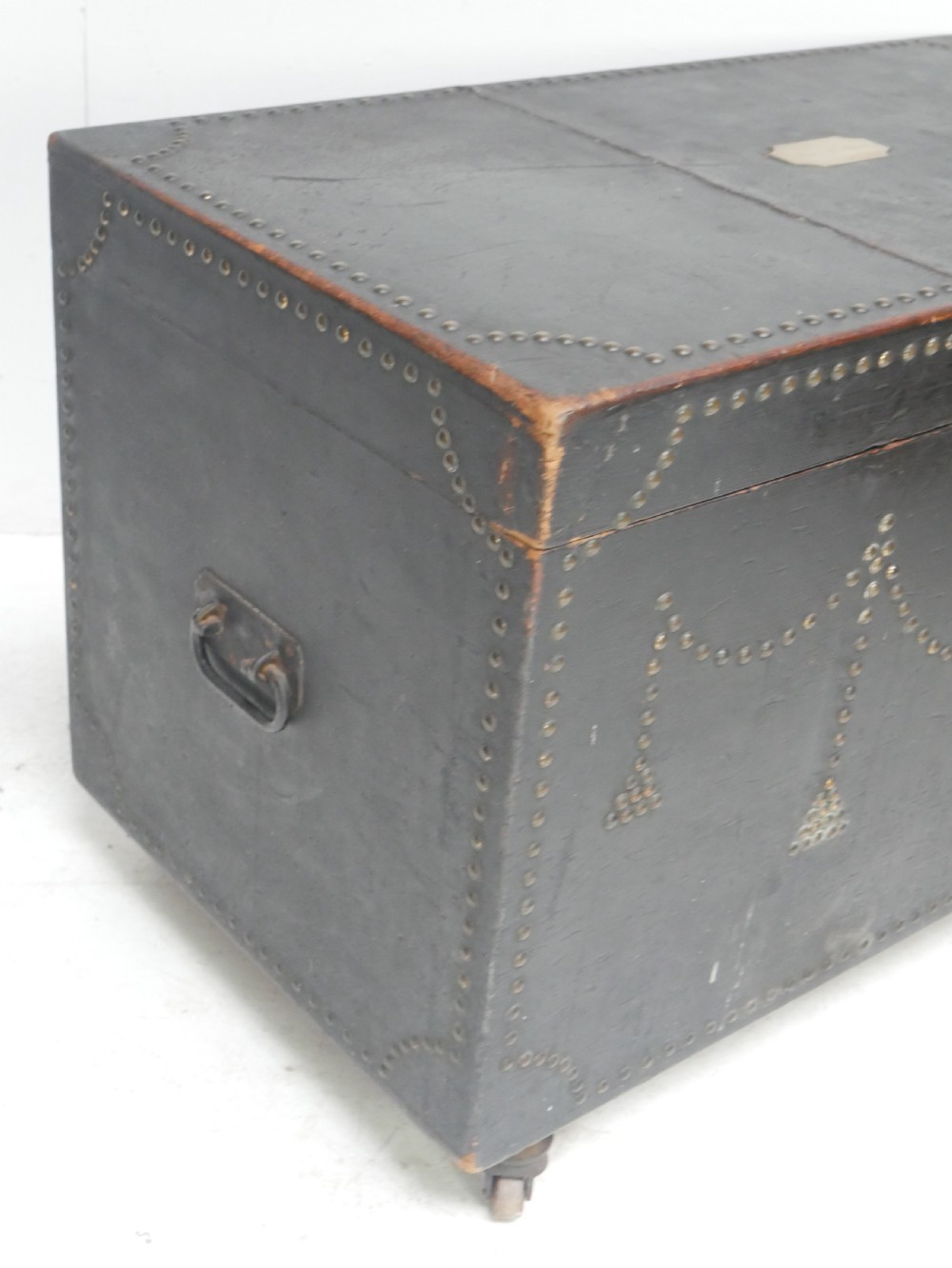 A black leather-covered storage trunk adorned with brass studding in festoon drape patterns, featuring side handles and castors. View VI