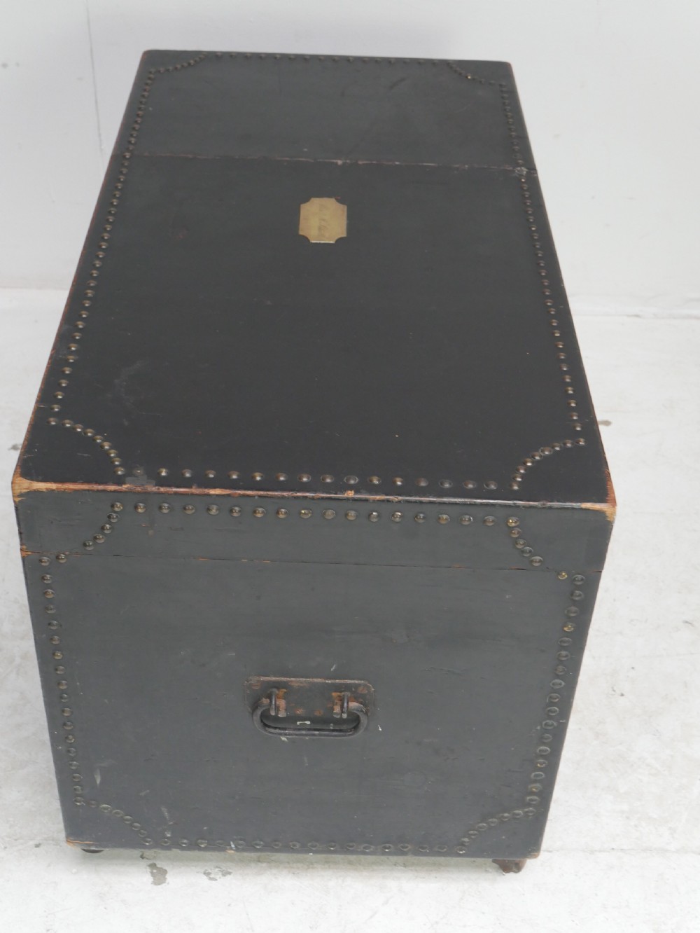 A black leather-covered storage trunk adorned with brass studding in festoon drape patterns, featuring side handles and castors. View VII
