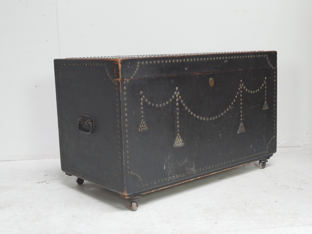 A black leather-covered storage trunk adorned with brass studding in festoon drape patterns, featuring side handles and castors. View XII