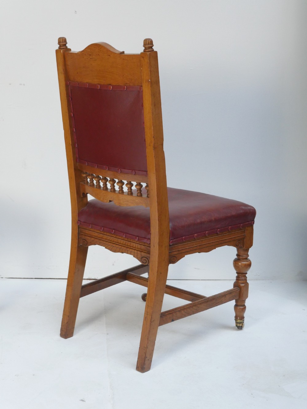 A pair of Victorian carved oak side chairs featuring reddish-brown leather seats and backrests secured with brass studs, set on turned front legs with castors. View V