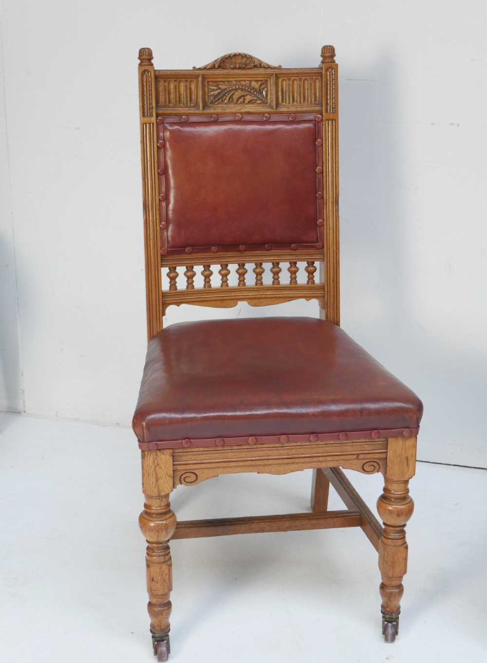 A pair of Victorian carved oak side chairs featuring reddish-brown leather seats and backrests secured with brass studs, set on turned front legs with castors. View IX