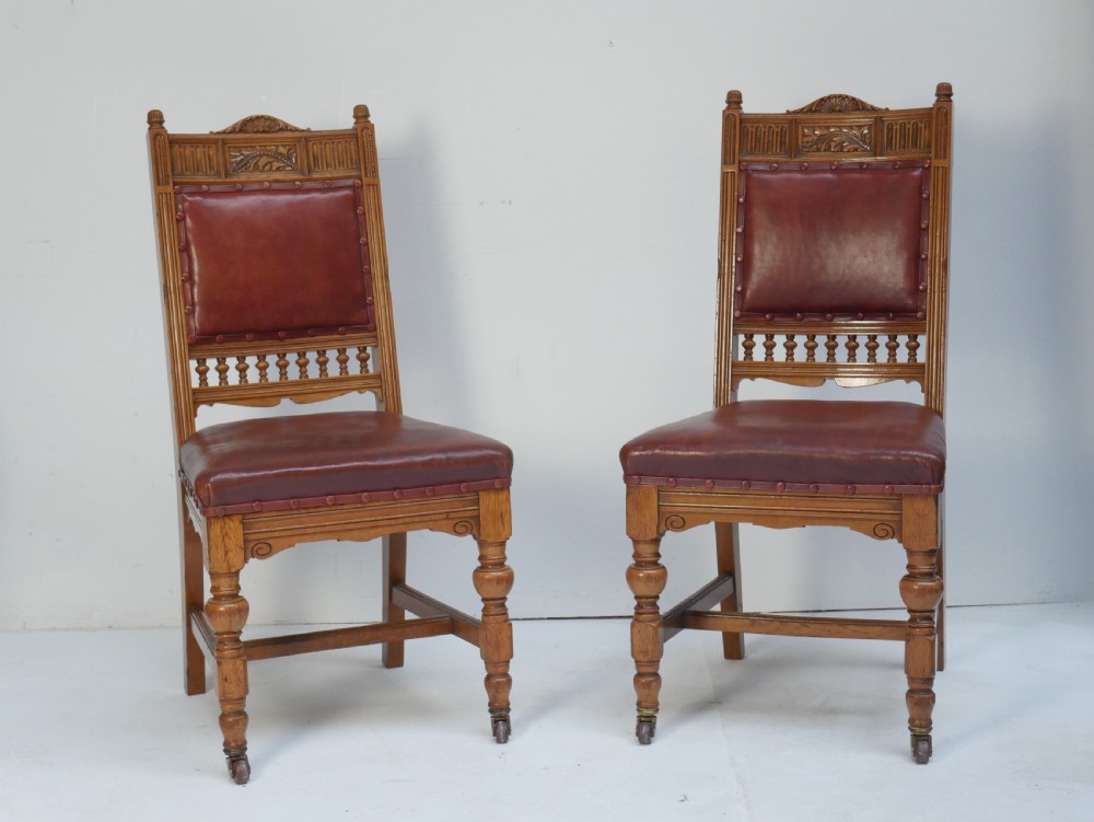 A pair of Victorian carved oak side chairs featuring reddish-brown leather seats and backrests secured with brass studs, set on turned front legs with castors. View X