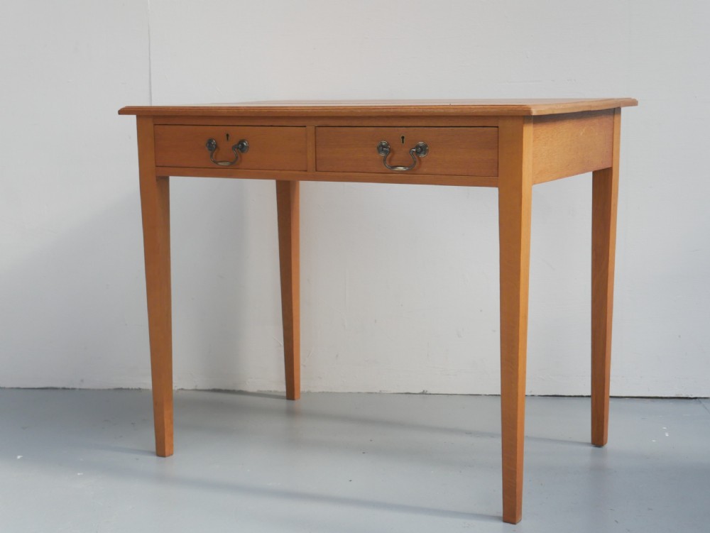 A golden oak writing table with two frieze drawers fitted with brass drop handles, raised on four square tapered legs. View II