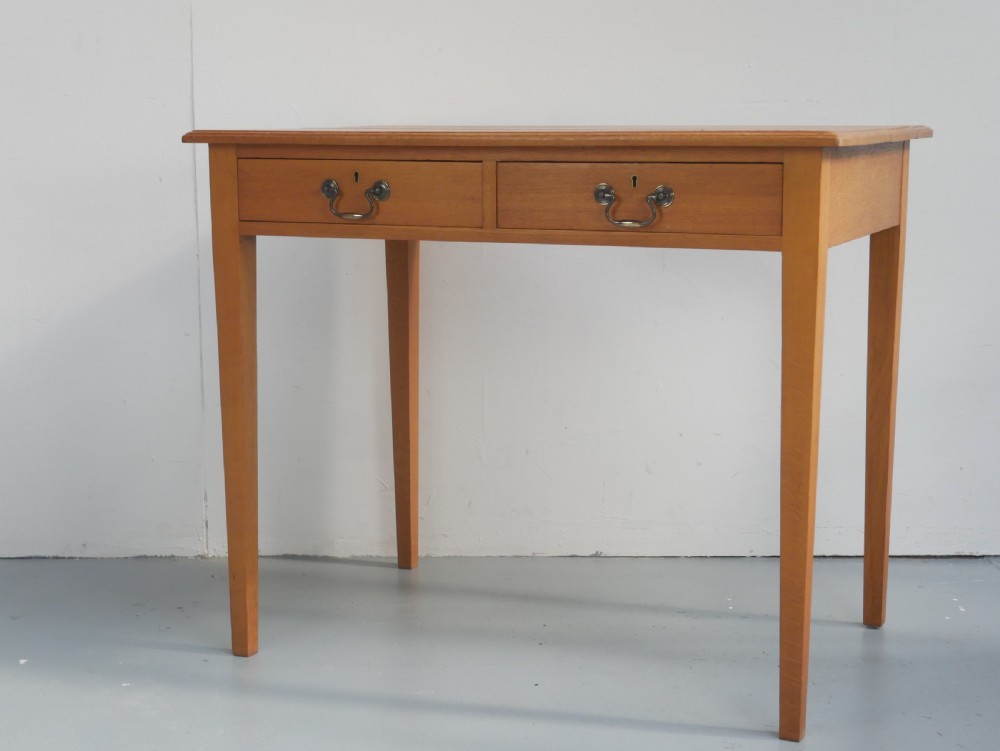A golden oak writing table with two frieze drawers fitted with brass drop handles, raised on four square tapered legs. View III