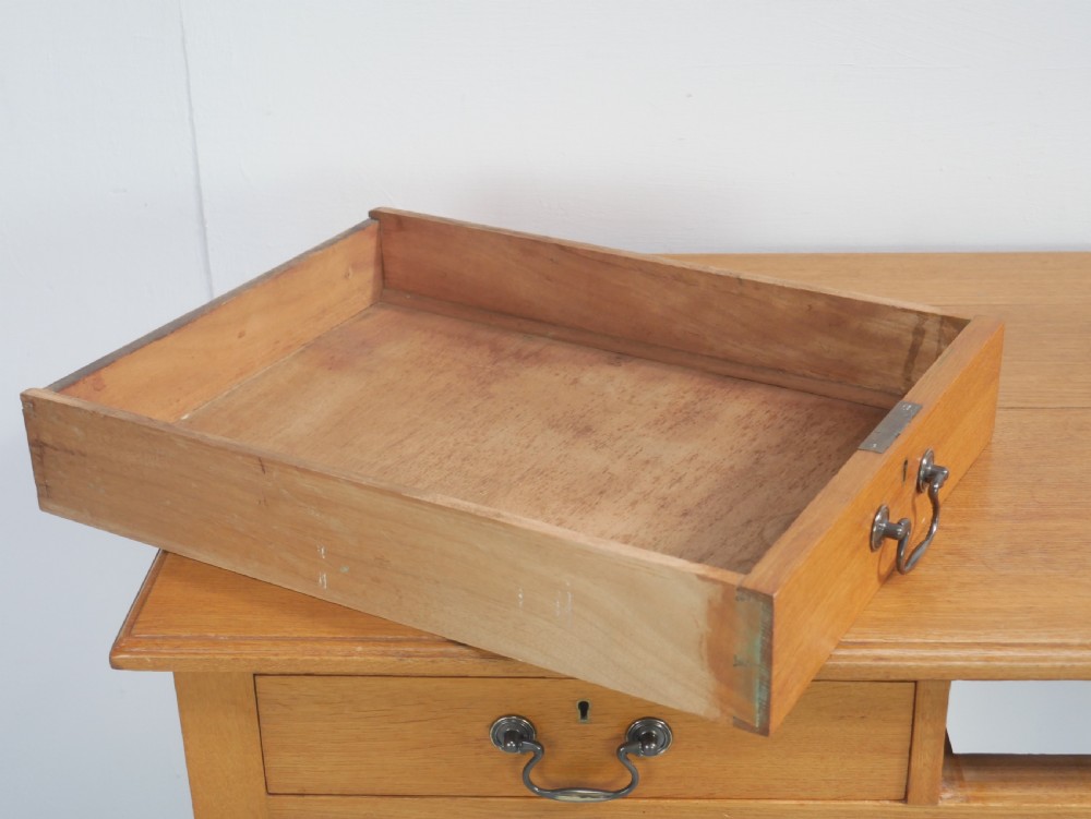 A golden oak writing table with two frieze drawers fitted with brass drop handles, raised on four square tapered legs. View VI