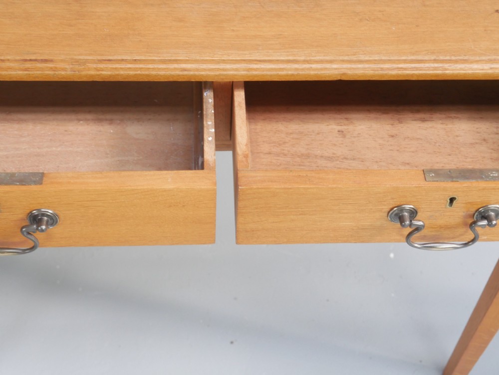 A golden oak writing table with two frieze drawers fitted with brass drop handles, raised on four square tapered legs. View VII