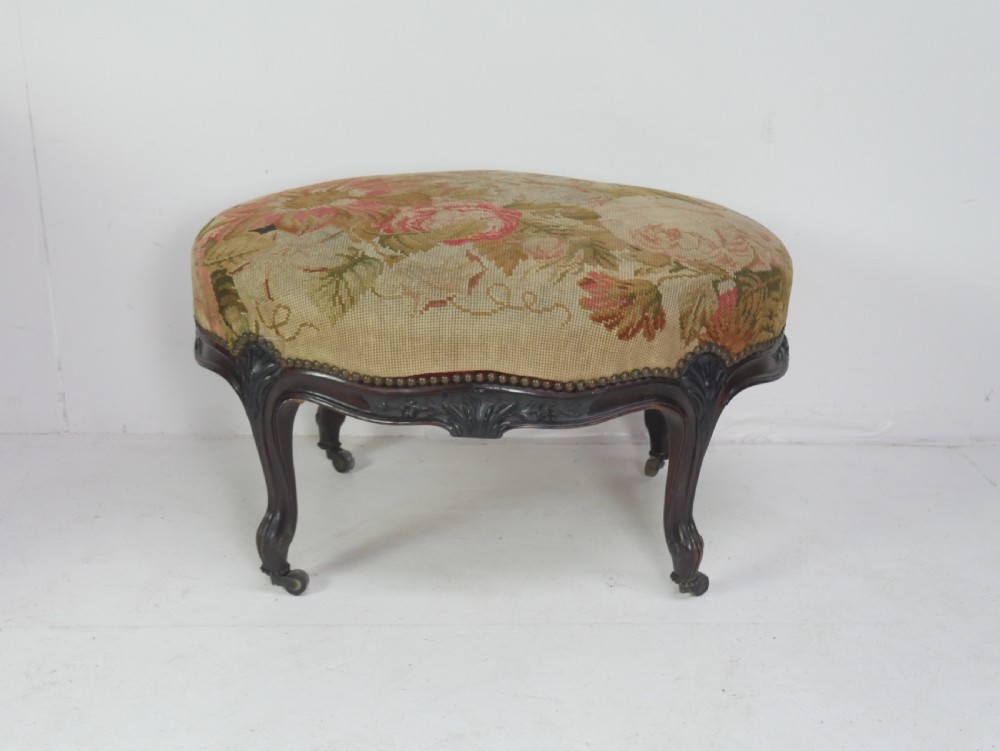 An oval upholstered stool featuring dark carved wooden cabriole legs with castors and a floral needlepoint fabric top.