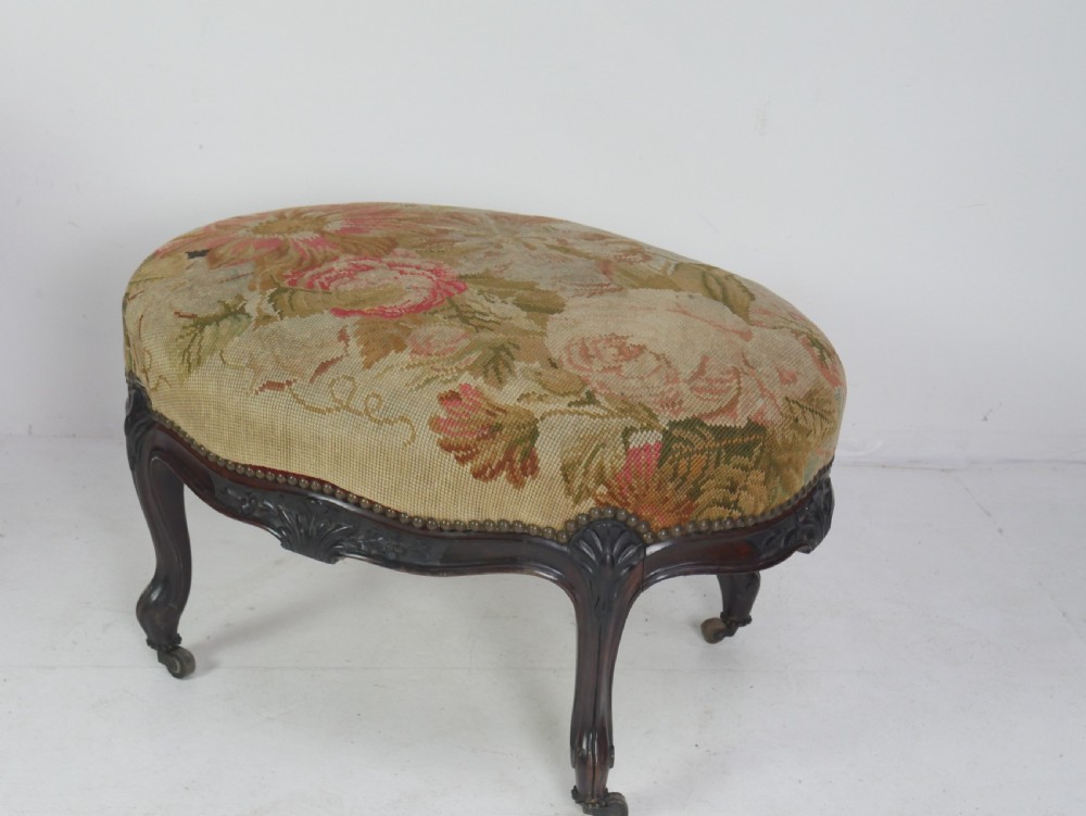 An oval upholstered stool featuring dark carved wooden cabriole legs with castors and a floral needlepoint fabric top. View II
