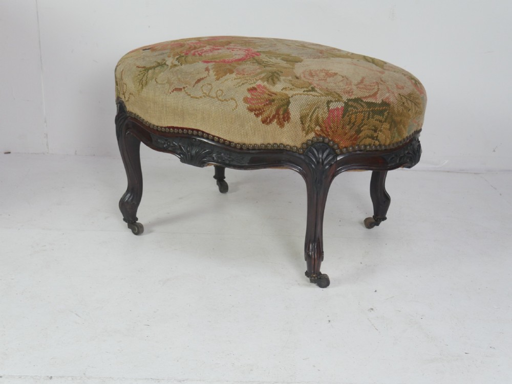 An oval upholstered stool featuring dark carved wooden cabriole legs with castors and a floral needlepoint fabric top. View VII