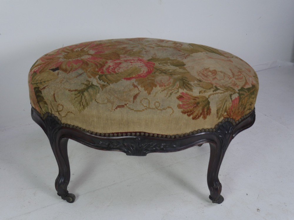 An oval upholstered stool featuring dark carved wooden cabriole legs with castors and a floral needlepoint fabric top. View VIII