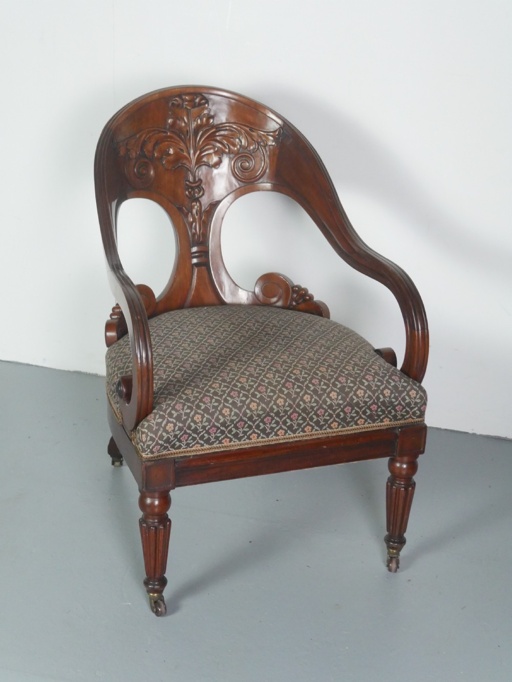 A dark mahogany Regency armchair with a carved leaf back splat, open scrolled arms, patterned upholstered seat, and turned front legs with brass castors.