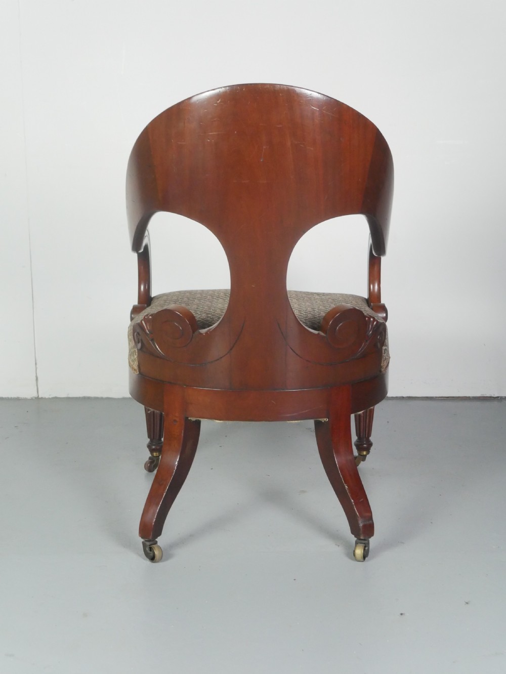 A dark mahogany Regency armchair with a carved leaf back splat, open scrolled arms, patterned upholstered seat, and turned front legs with brass castors. View V