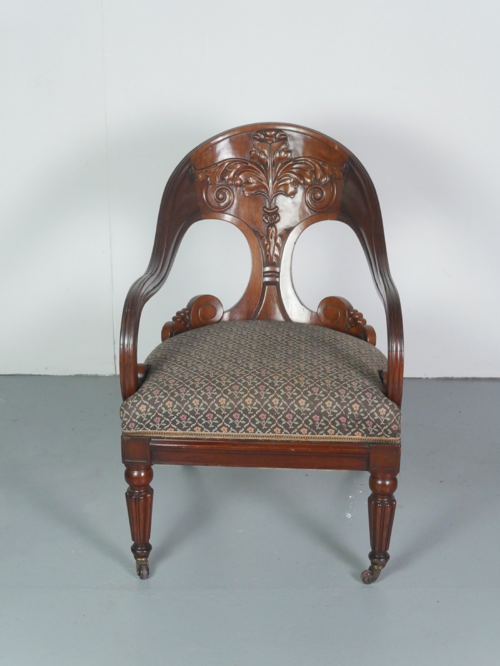 A dark mahogany Regency armchair with a carved leaf back splat, open scrolled arms, patterned upholstered seat, and turned front legs with brass castors. View X