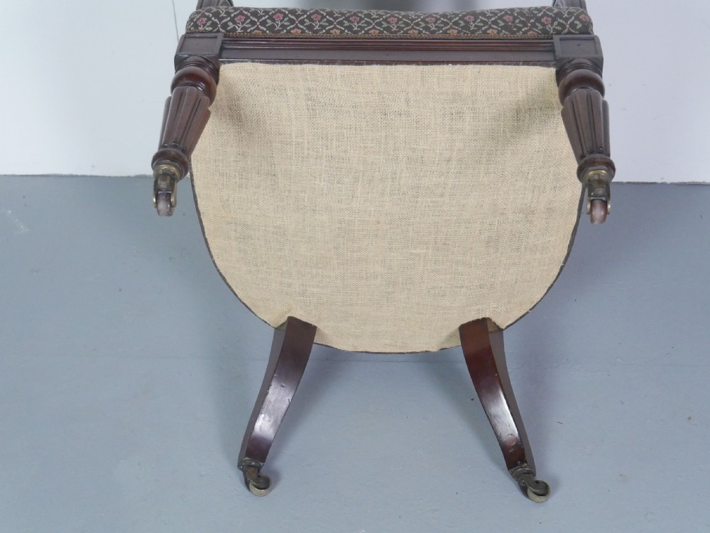 A dark mahogany Regency armchair with a carved leaf back splat, open scrolled arms, patterned upholstered seat, and turned front legs with brass castors. View XI