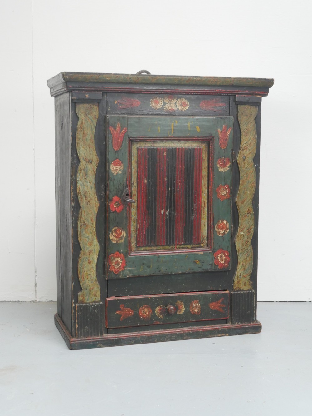 An antique dark green Swedish wall cupboard featuring hand-painted floral motifs, a central panel door, and a single lower drawer.