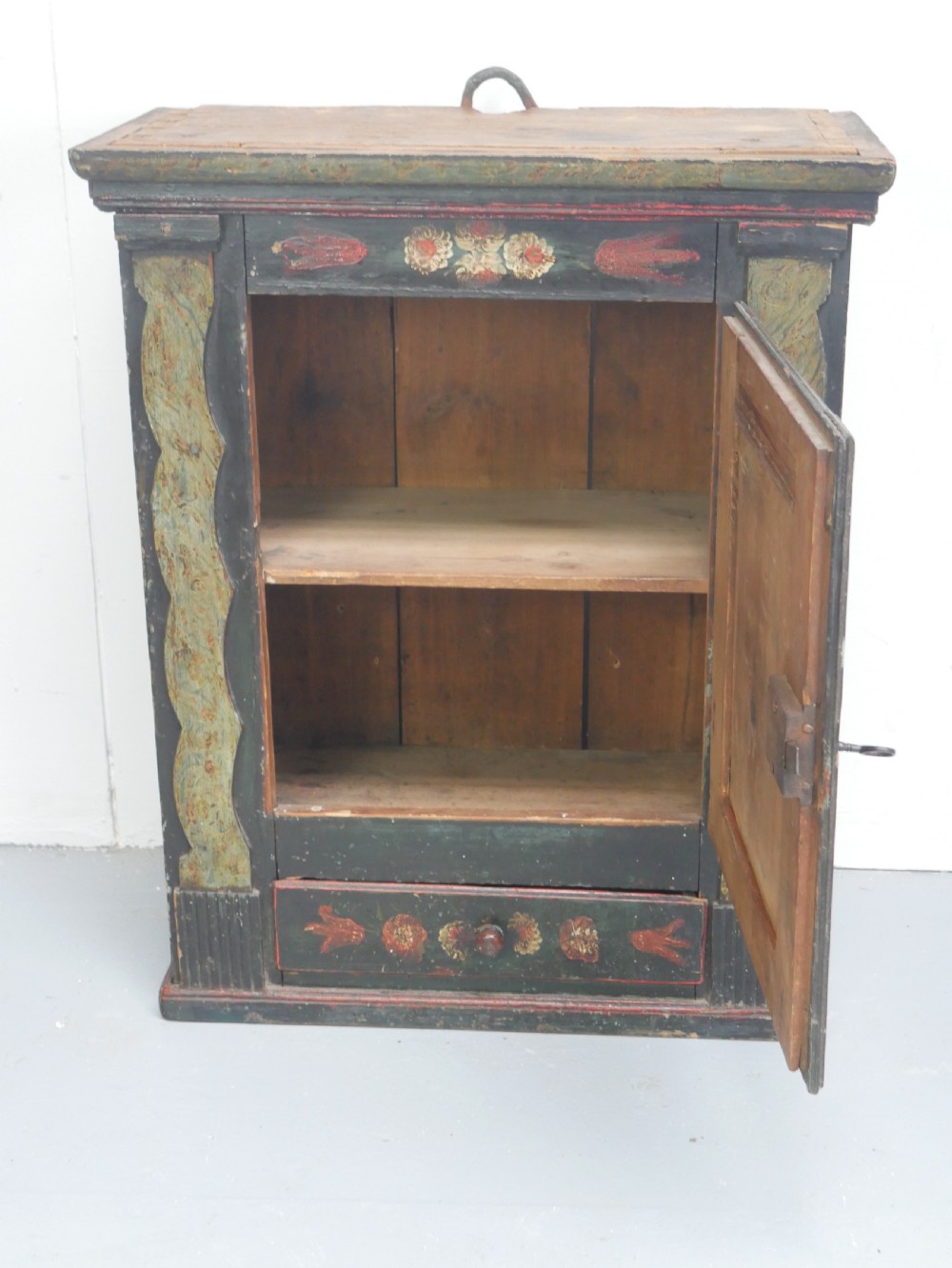 An antique dark green Swedish wall cupboard featuring hand-painted floral motifs, a central panel door, and a single lower drawer. View II