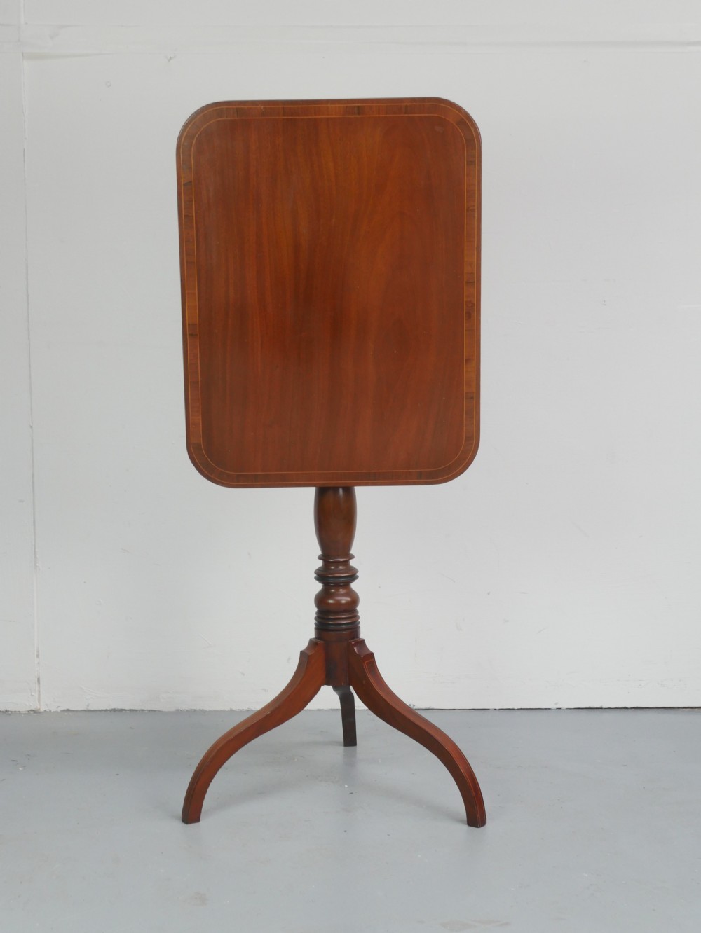 An antique mahogany tilt-top table with a rectangular inlaid tabletop raised vertically on a turned central pedestal and three curved legs.