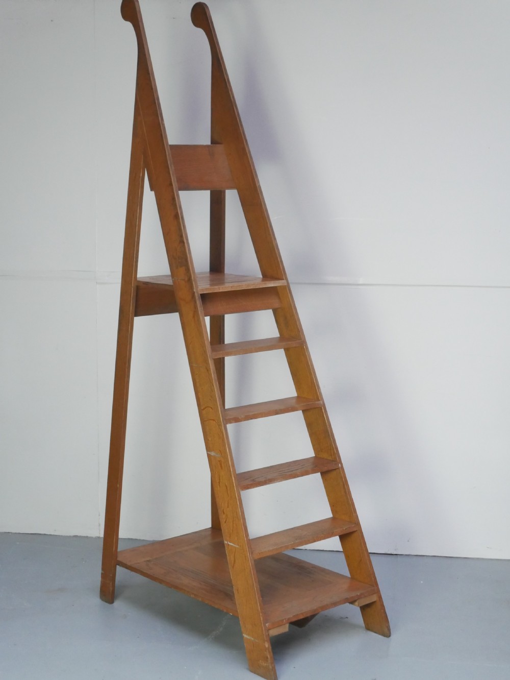 A tall freestanding oak library step ladder featuring multiple wide treads, shaped handholds at the top, and a warm natural wood finish.
