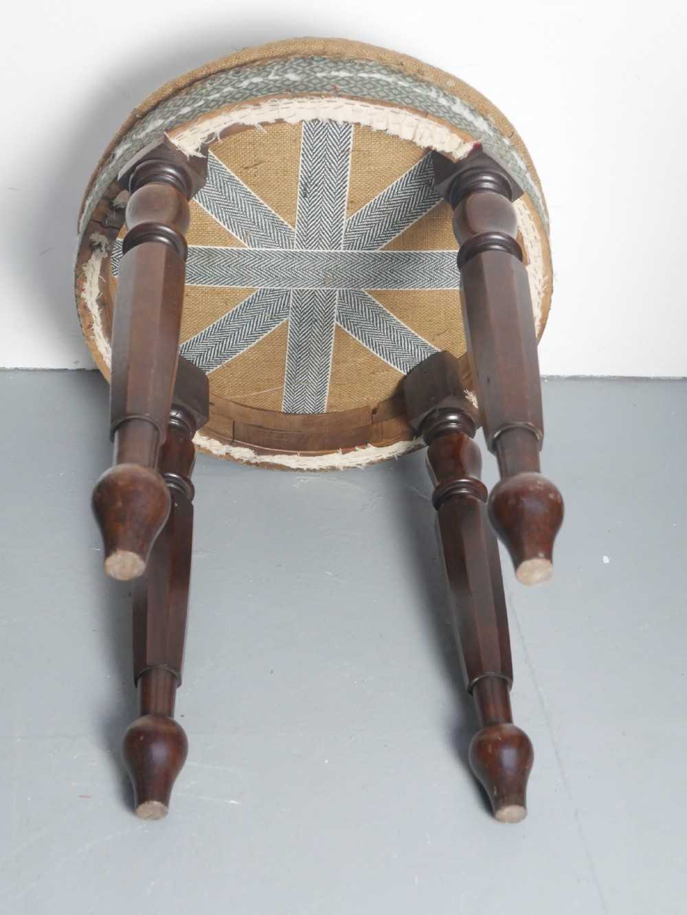 A circular stool with four dark turned timber legs and a padded circular top showing visible upholstery layers. View VI