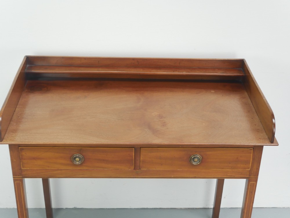 An antique mahogany writing table featuring a low gallery shelf, two drawers with circular brass pull handles, line inlay details, and four square tapered legs. View II
