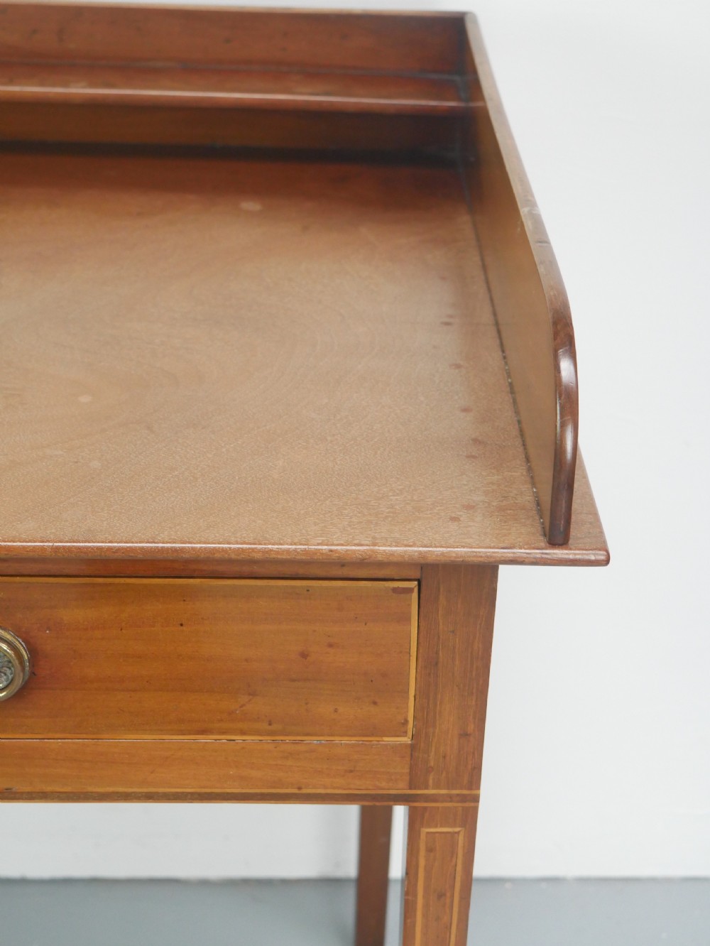 An antique mahogany writing table featuring a low gallery shelf, two drawers with circular brass pull handles, line inlay details, and four square tapered legs. View VI