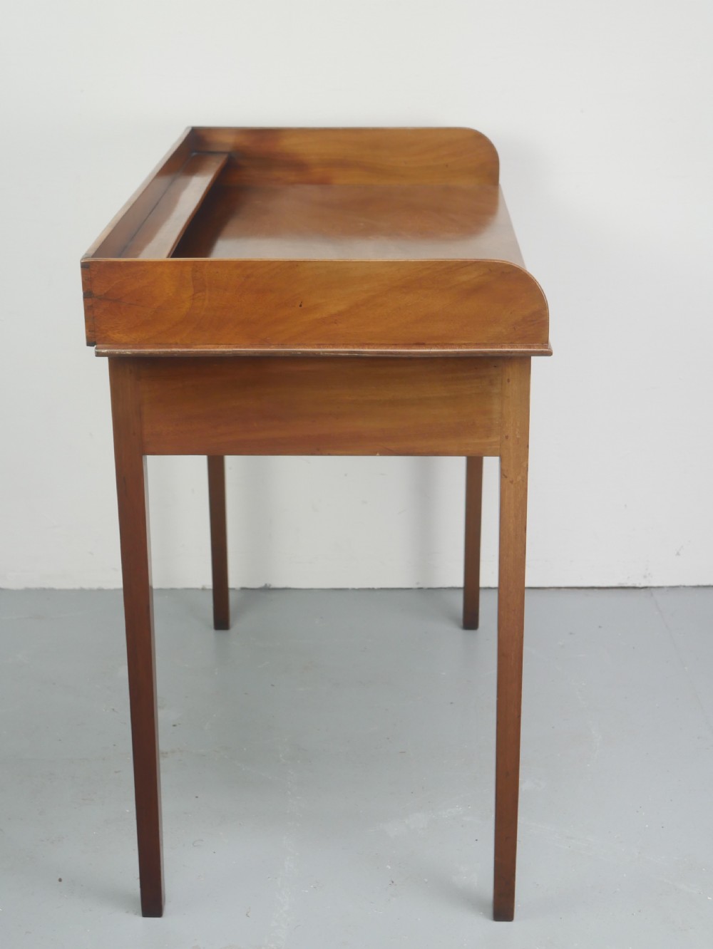 An antique mahogany writing table featuring a low gallery shelf, two drawers with circular brass pull handles, line inlay details, and four square tapered legs. View VII