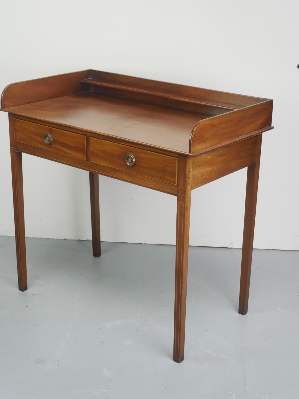 An antique mahogany writing table featuring a low gallery shelf, two drawers with circular brass pull handles, line inlay details, and four square tapered legs. View X