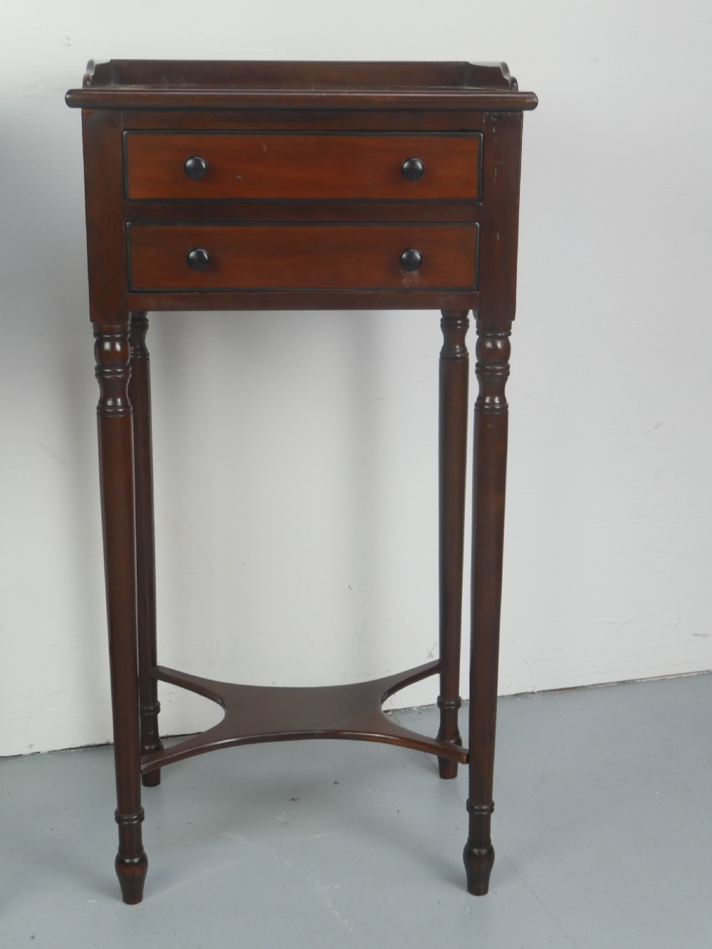 A pair of tall slim dark mahogany bedside tables featuring tray tops, two drawers with black knobs, turned legs, and curved X-frame stretchers. View IV