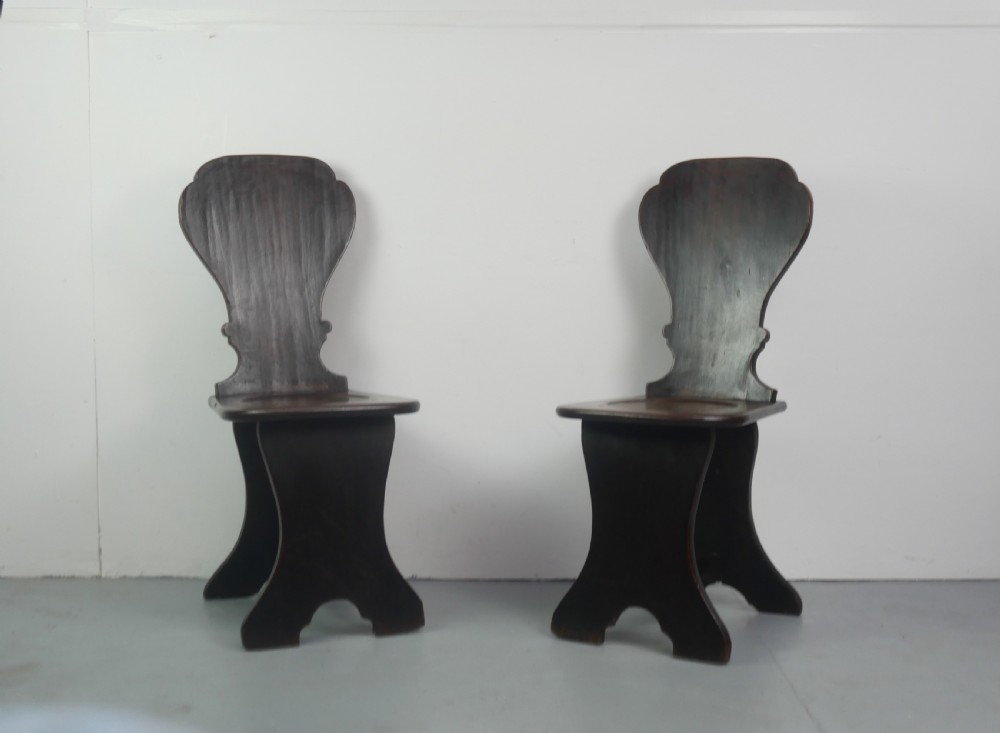 A pair of dark brown 18th-century oak sgabello hall chairs with shaped backrests and trestle bases set against a plain white background.