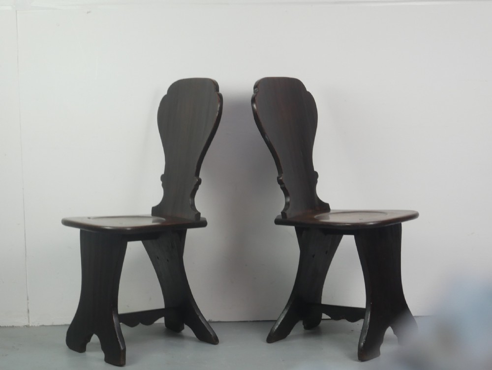 A pair of dark brown 18th-century oak sgabello hall chairs with shaped backrests and trestle bases set against a plain white background. View II