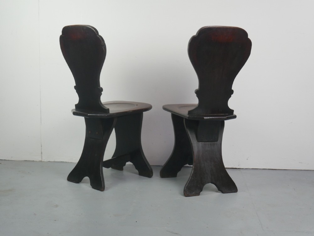 A pair of dark brown 18th-century oak sgabello hall chairs with shaped backrests and trestle bases set against a plain white background. View IX