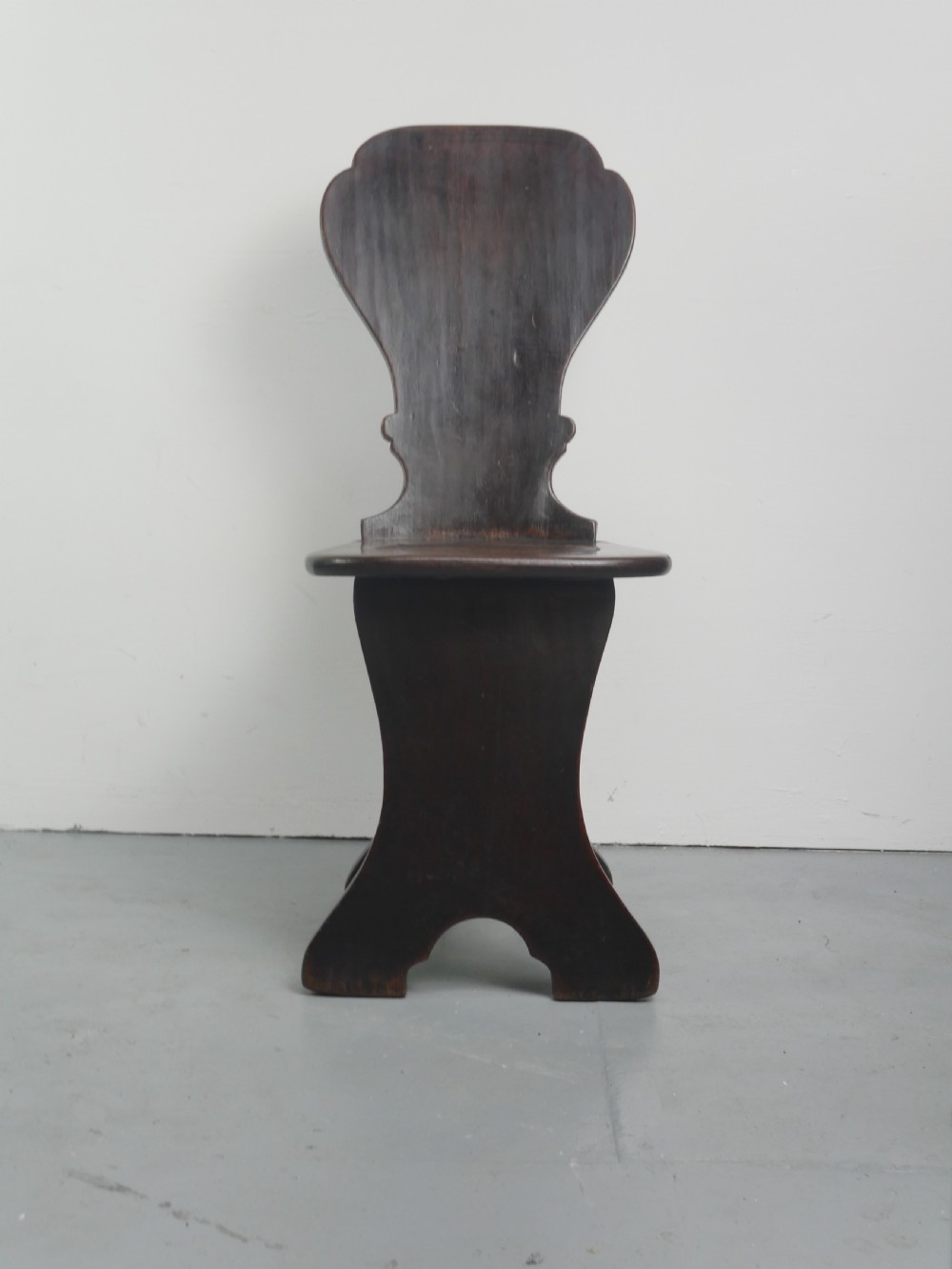 A pair of dark brown 18th-century oak sgabello hall chairs with shaped backrests and trestle bases set against a plain white background. View X