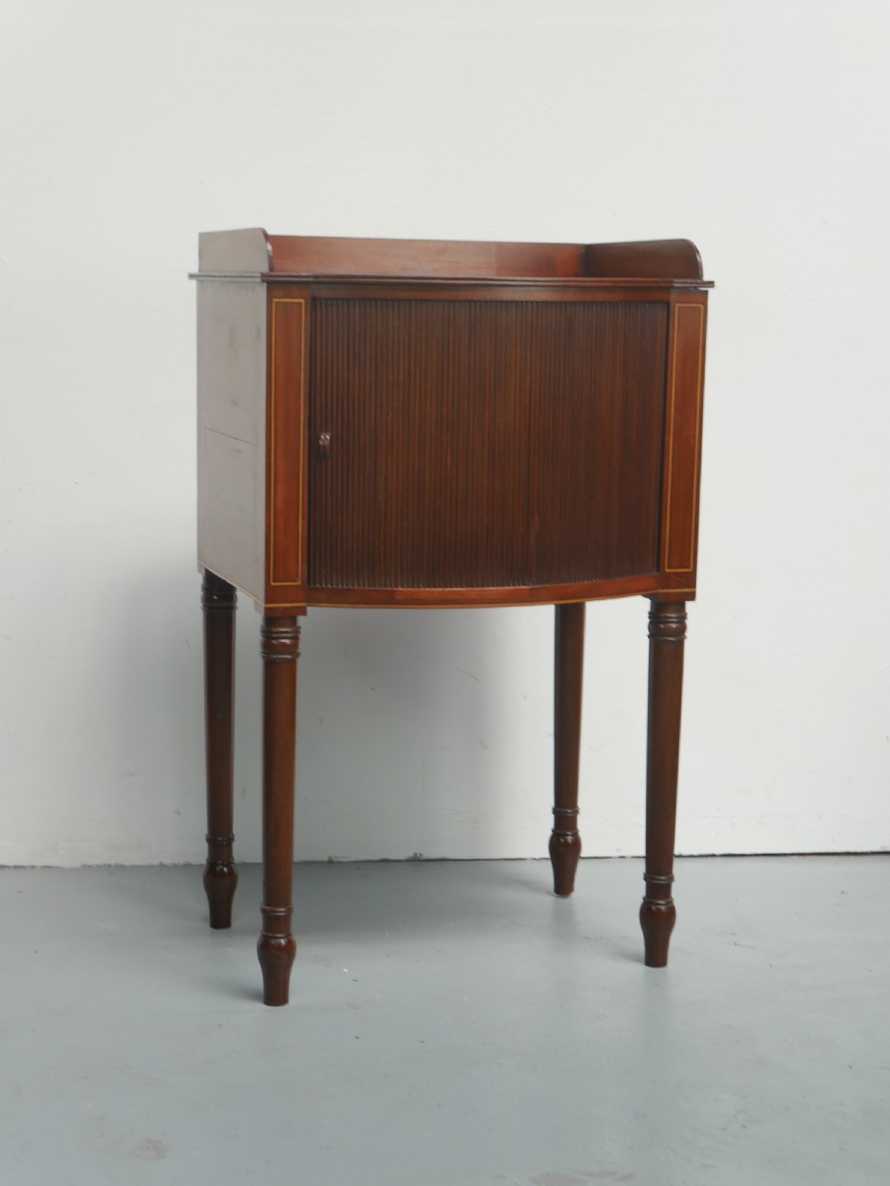 A Regency mahogany bedside cupboard featuring a reeded tambour door, fine boxwood stringing inlay, a raised tray top gallery, and slender turned legs.