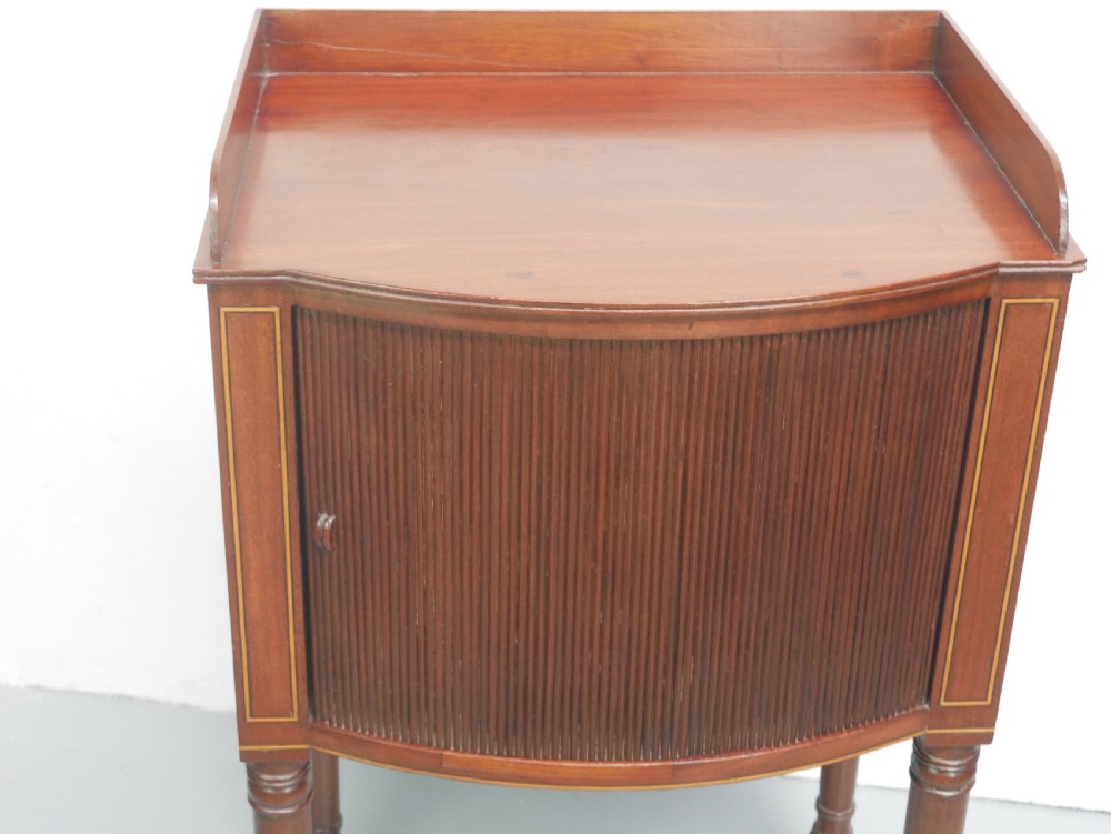 A Regency mahogany bedside cupboard featuring a reeded tambour door, fine boxwood stringing inlay, a raised tray top gallery, and slender turned legs. View II