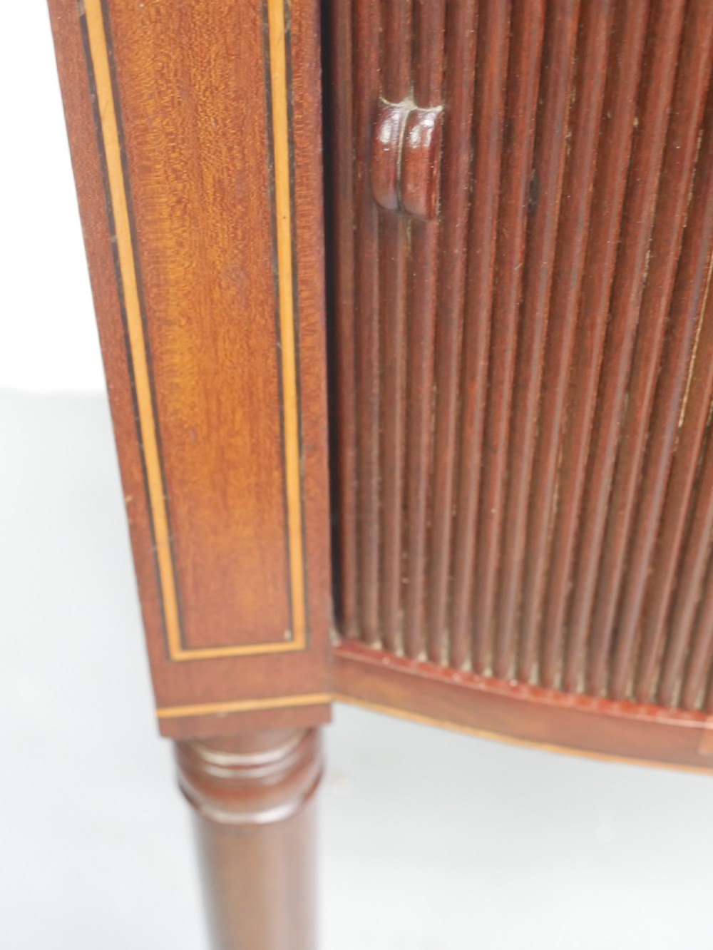 A Regency mahogany bedside cupboard featuring a reeded tambour door, fine boxwood stringing inlay, a raised tray top gallery, and slender turned legs. View V
