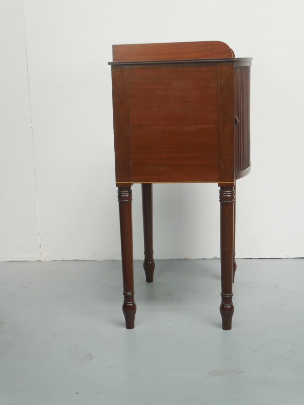 A Regency mahogany bedside cupboard featuring a reeded tambour door, fine boxwood stringing inlay, a raised tray top gallery, and slender turned legs. View VI