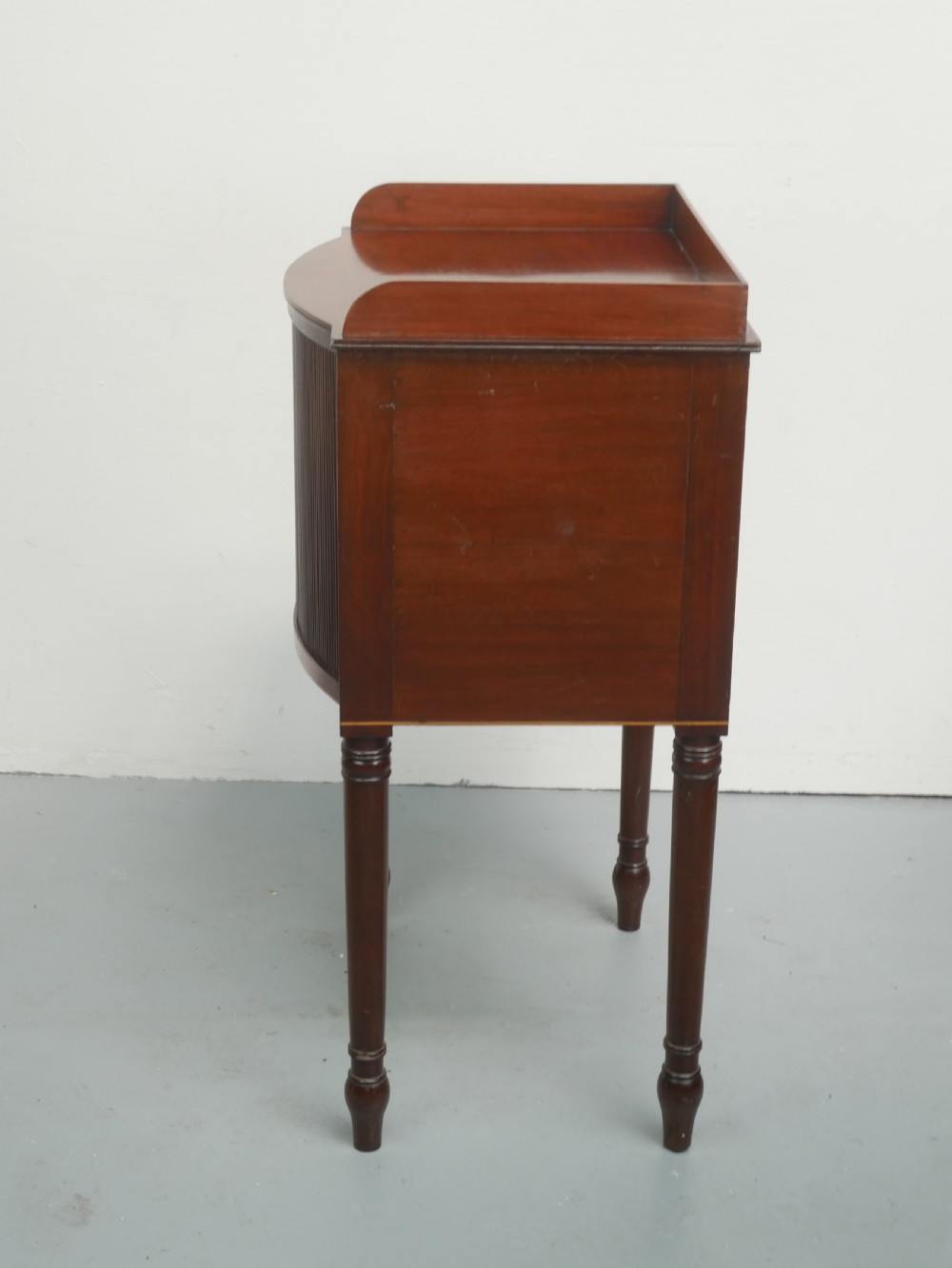 A Regency mahogany bedside cupboard featuring a reeded tambour door, fine boxwood stringing inlay, a raised tray top gallery, and slender turned legs. View VIII