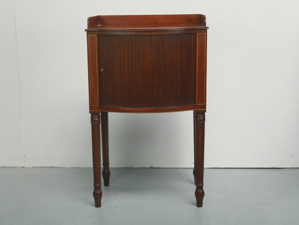 A Regency mahogany bedside cupboard featuring a reeded tambour door, fine boxwood stringing inlay, a raised tray top gallery, and slender turned legs. View X