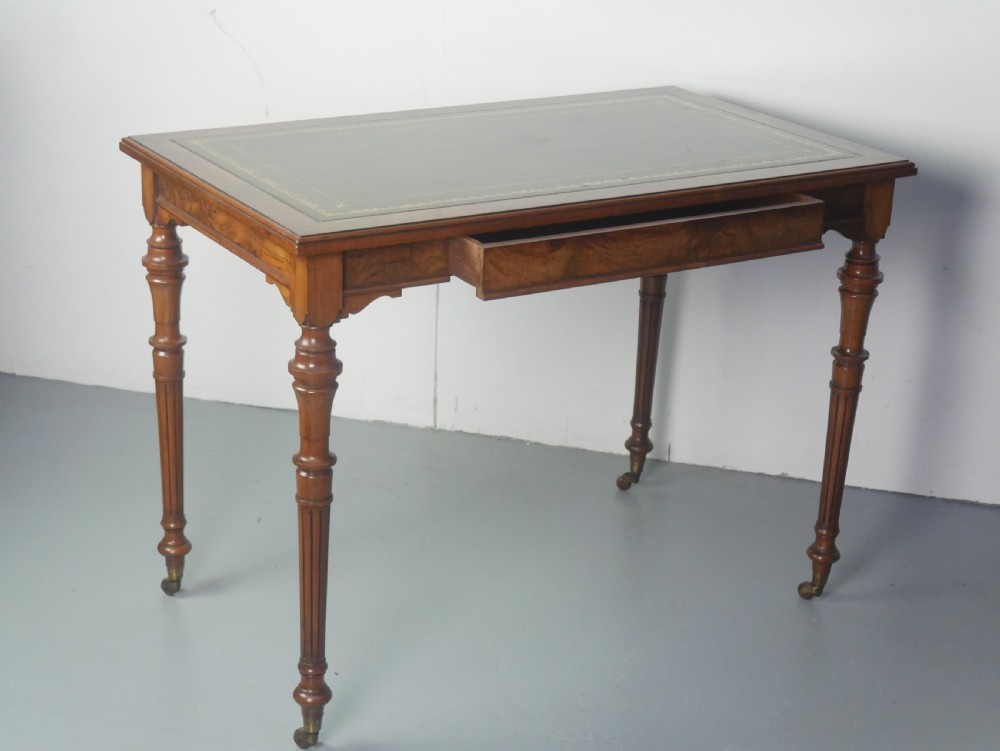 A light walnut writing table featuring a green leather inset top with gilt tooling, a single frieze drawer, and turned fluted legs on brass castors.