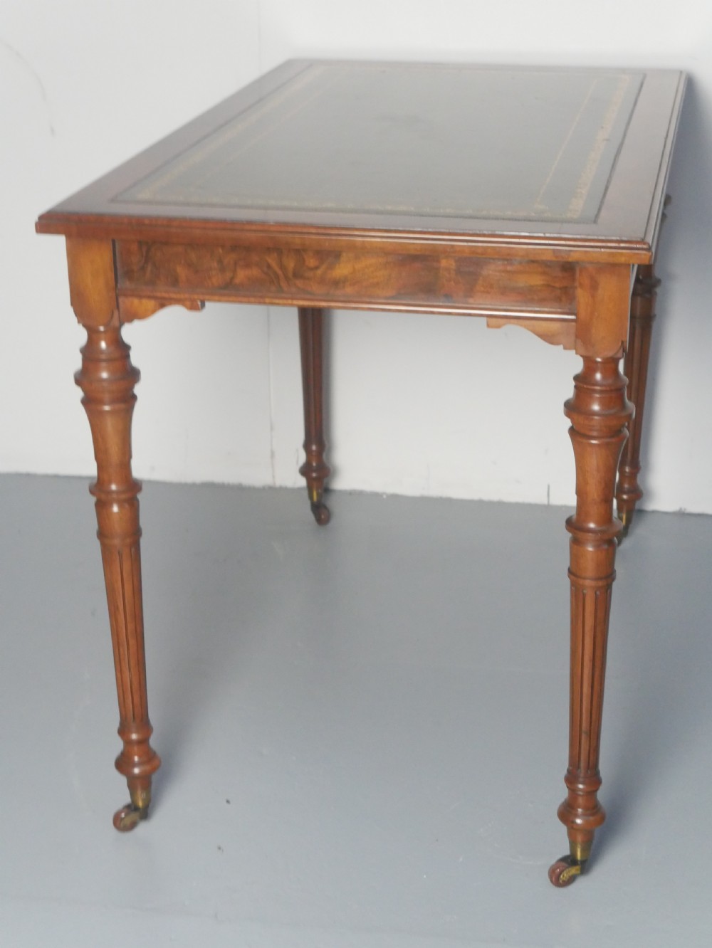 A light walnut writing table featuring a green leather inset top with gilt tooling, a single frieze drawer, and turned fluted legs on brass castors. View III