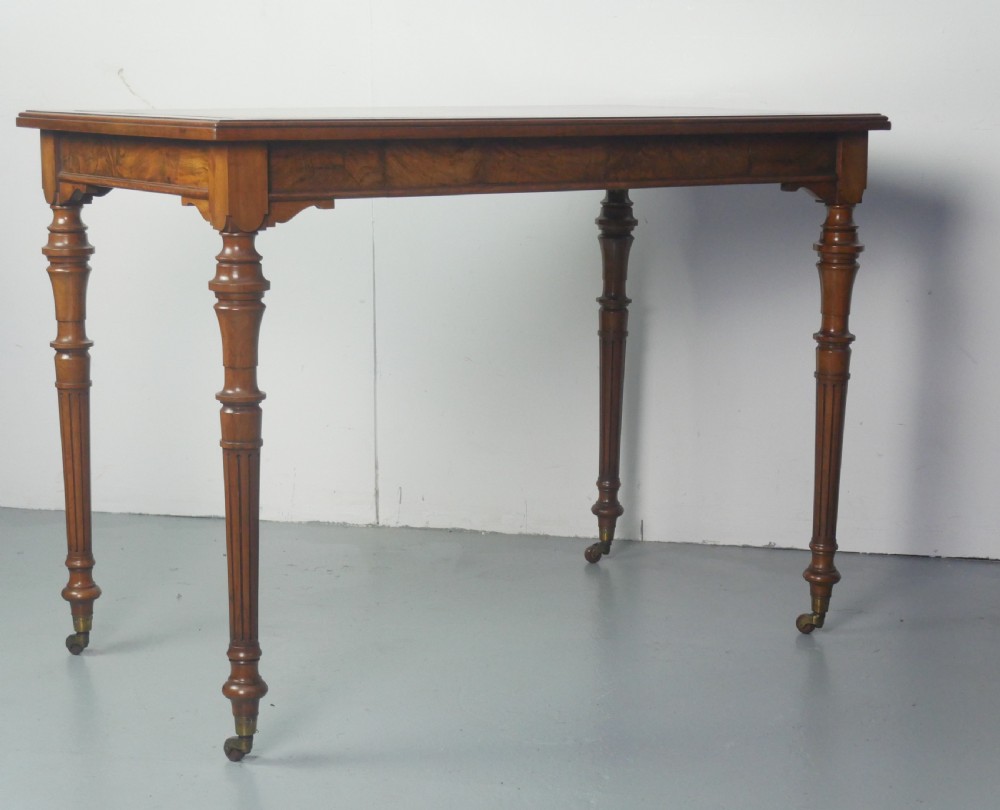 A light walnut writing table featuring a green leather inset top with gilt tooling, a single frieze drawer, and turned fluted legs on brass castors. View IX