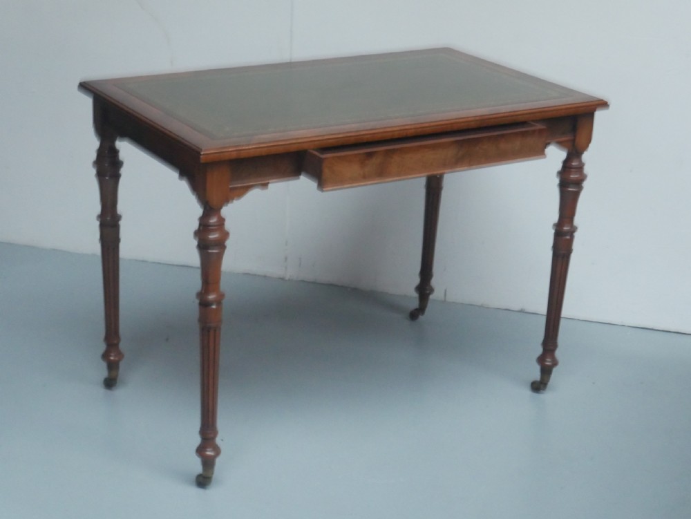 A light walnut writing table featuring a green leather inset top with gilt tooling, a single frieze drawer, and turned fluted legs on brass castors. View X
