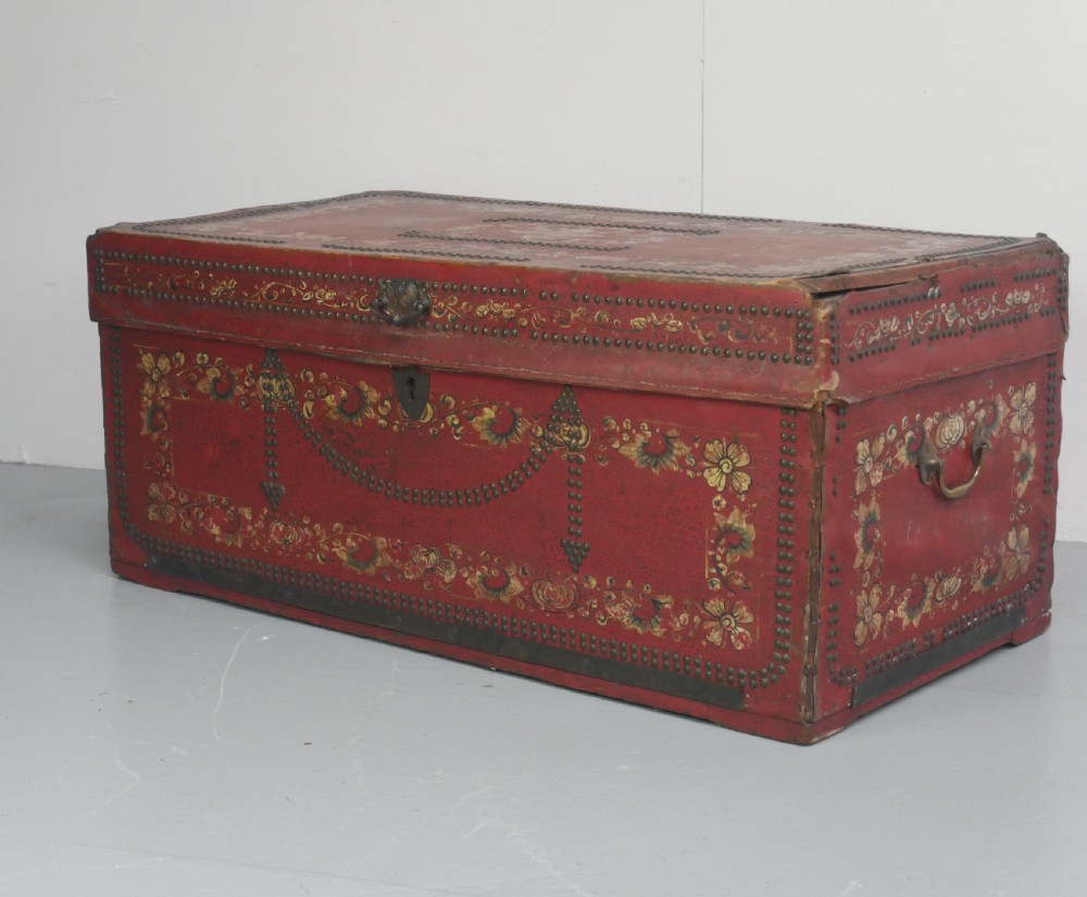 A rectangular red leather China trade trunk decorated with brass studs, brass side handles, and hand-painted gold floral motifs.