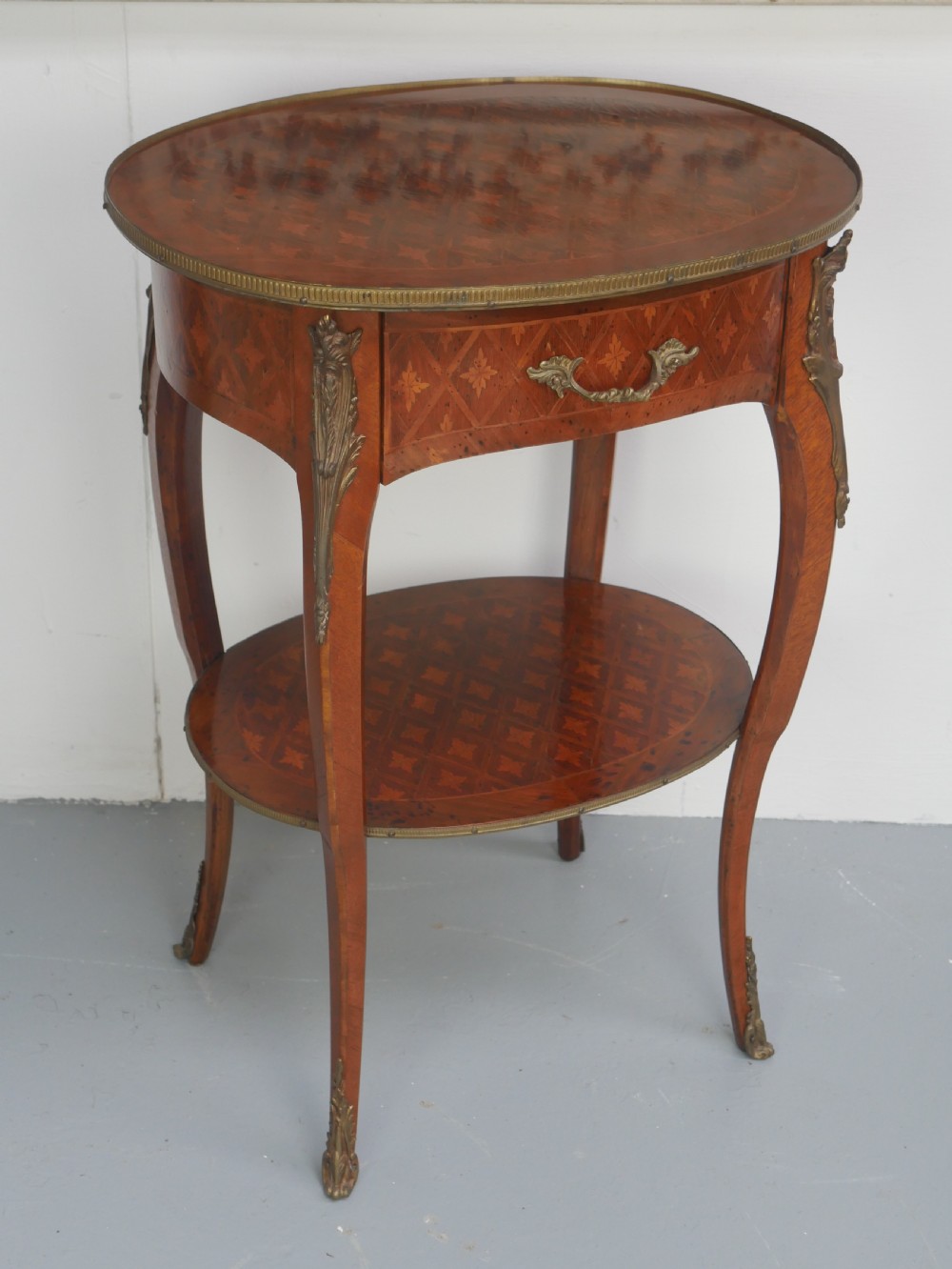 An oval Louis XV style wooden centre table featuring geometric parquetry inlay, a single drawer, a lower tier, cabriole legs, and ornate gilt metal mounts.